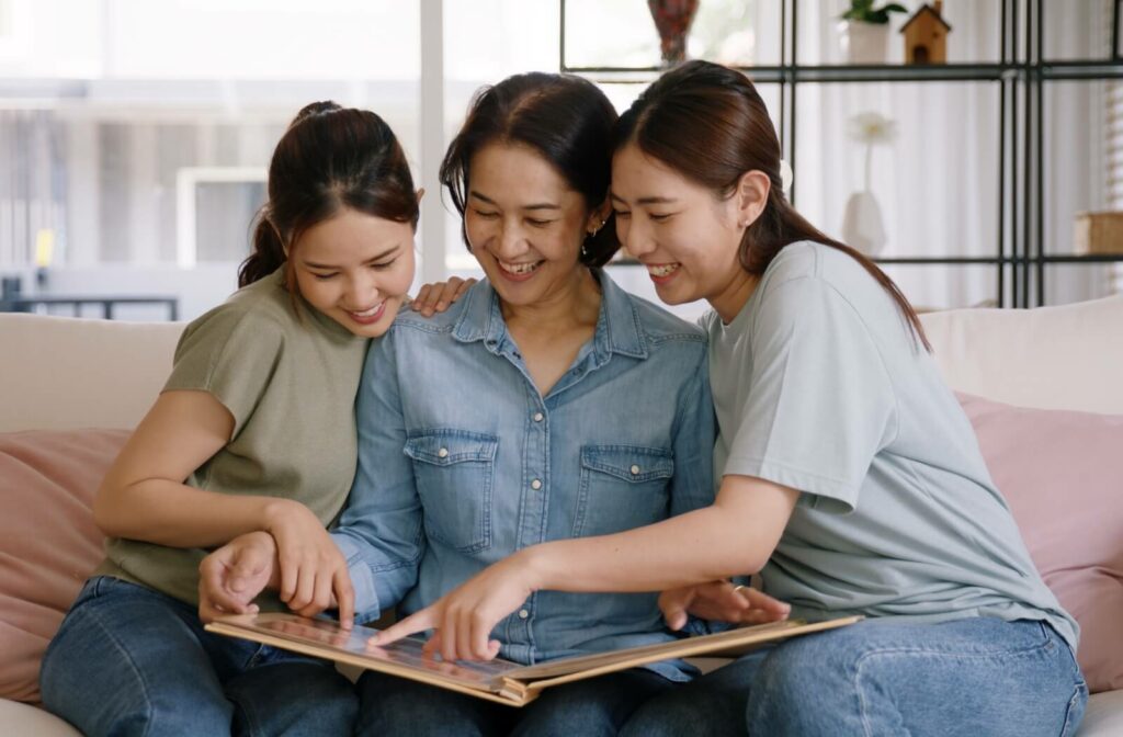 A senior holds a photo album on their lap, reminiscing with their children on either side of them as they point at pictures
