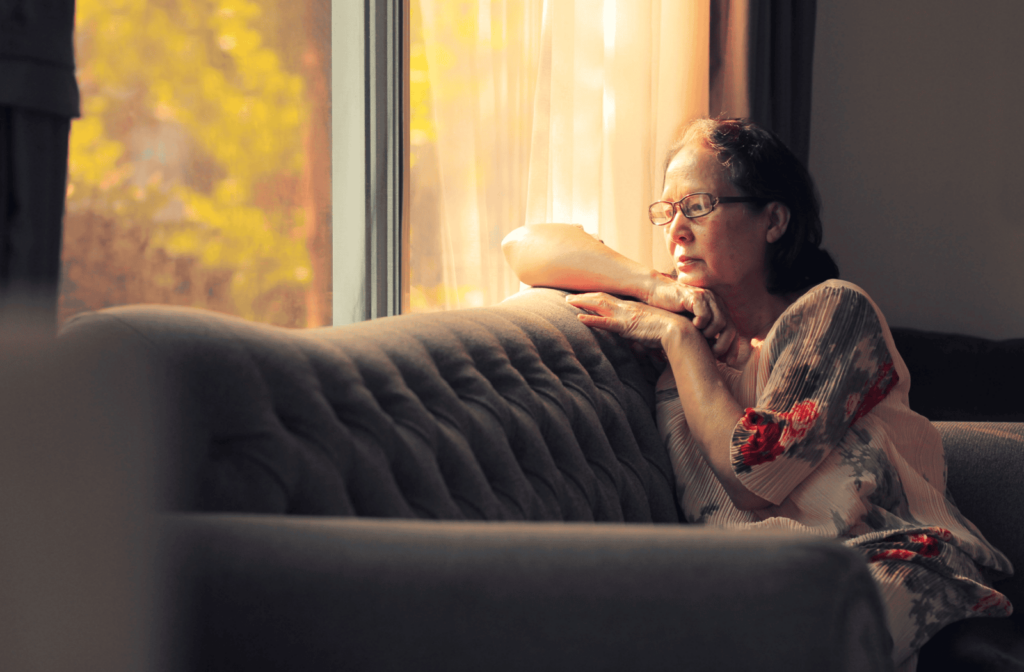 A senior person sits on their couch alone, staring out the window.