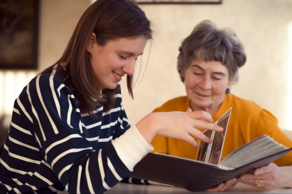 A senior and their grandchild sit at a dining room table, looking through a photo album together.