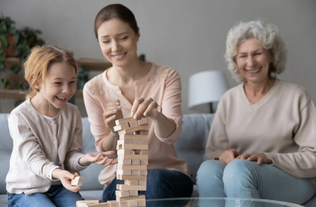 An older adult smiling and sitting on the couch while their grandchild and adult child assemble a block tower during a visit in skilled nursing.