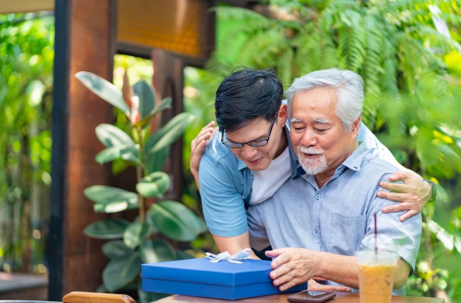 An adult surprises their older parent with a gift wrapped in a blue box while meeting up for boba in a public park