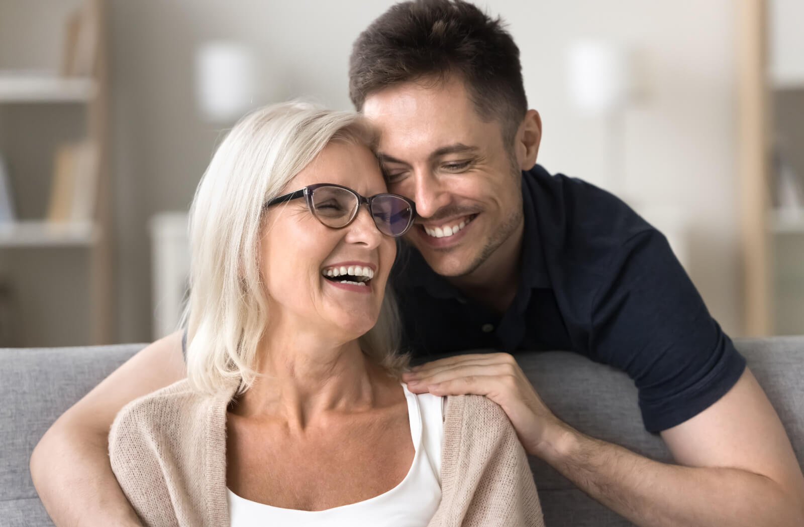 An older adult smiling while their adult child hugs them from behind during a visit in skilled nursing.