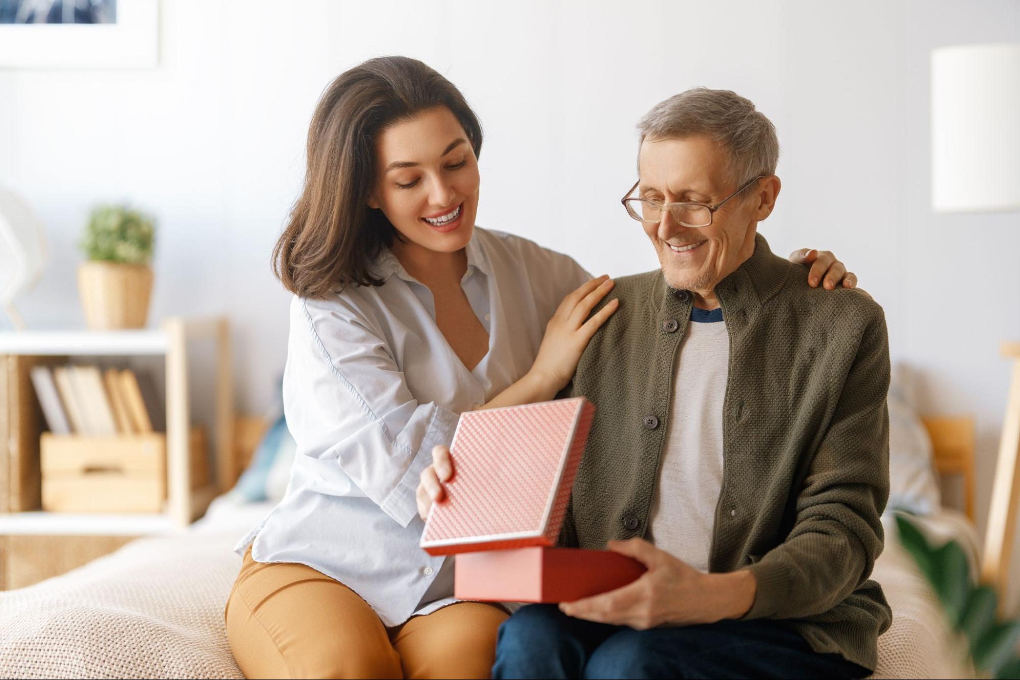 A senior and their adult child sit side by side on a bed. The senior is smiling and opening a gift from their child, while their child rests their hands affectionately on their parent's shoulders.