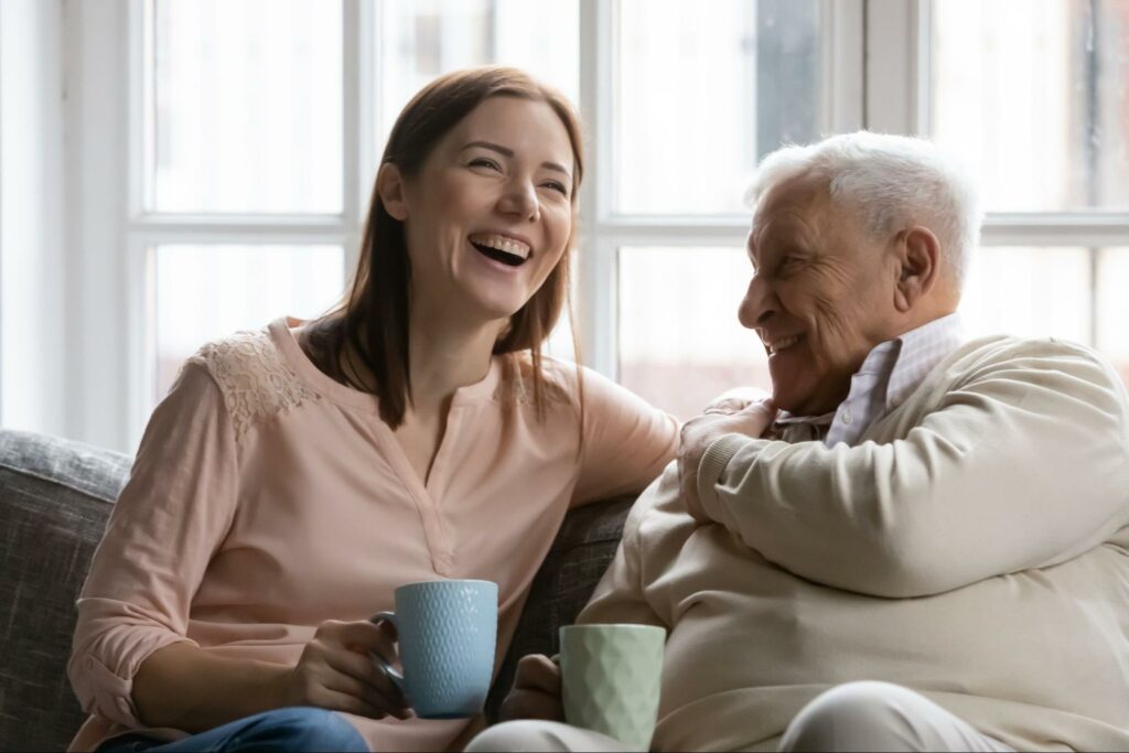 A woman and her senior father drinking tea and laughing during a visit to memory care.