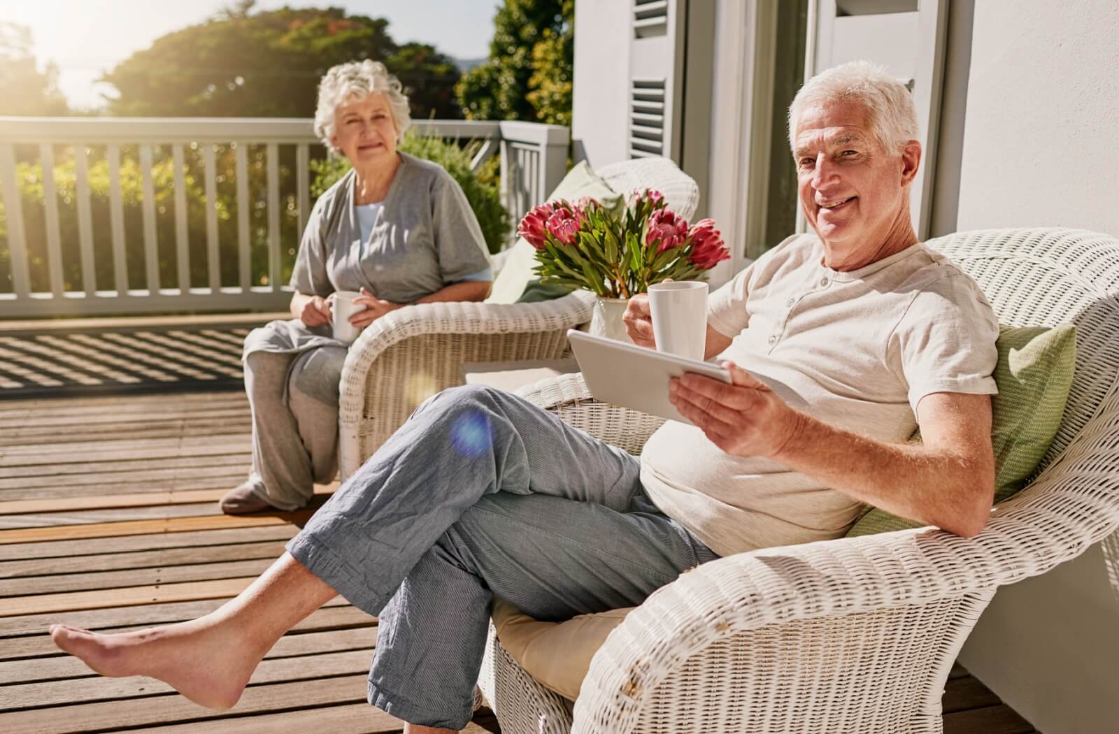Two older adults relaxing on a sunny porch with coffee and a tablet surrounded by flowers and outdoor furniture