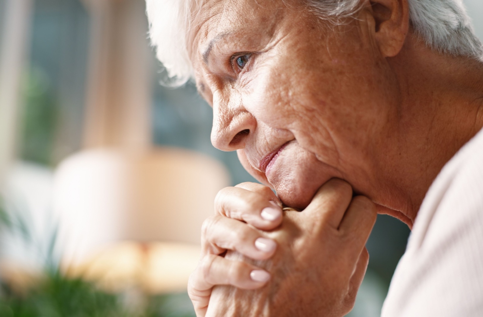 An older adult sits indoors near a bright window with their hands clasped under their chin.