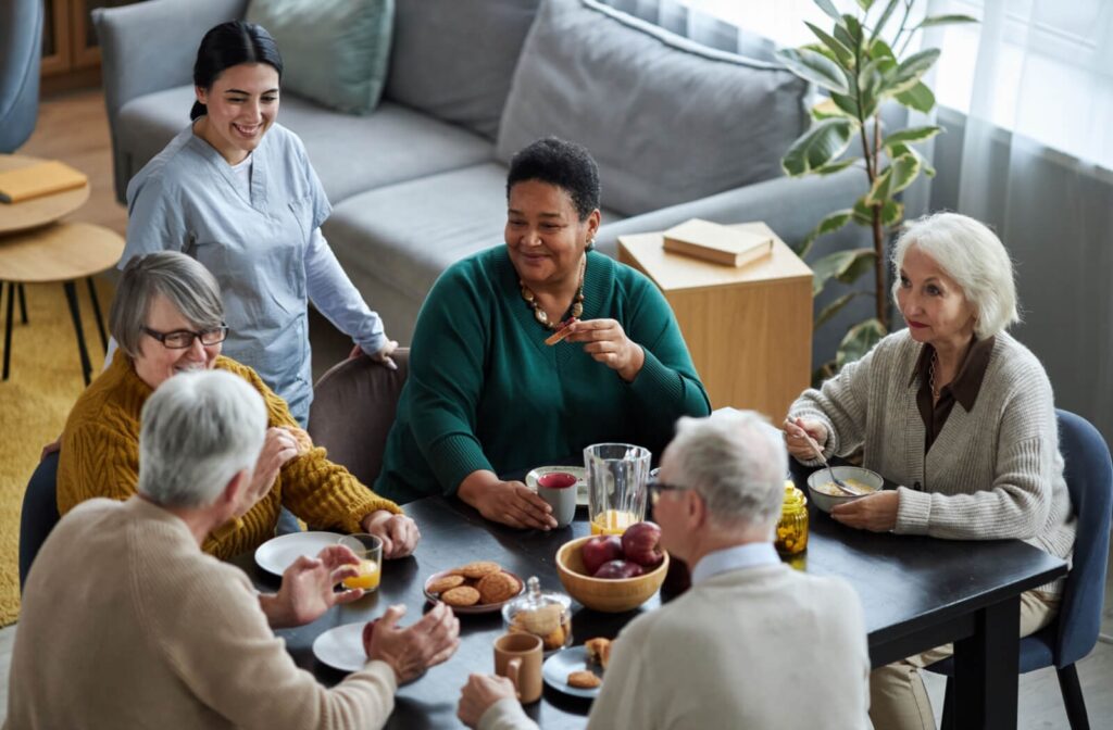 A group of seniors sits around a table, eating and talking together, while a smiling caregiver checks in.